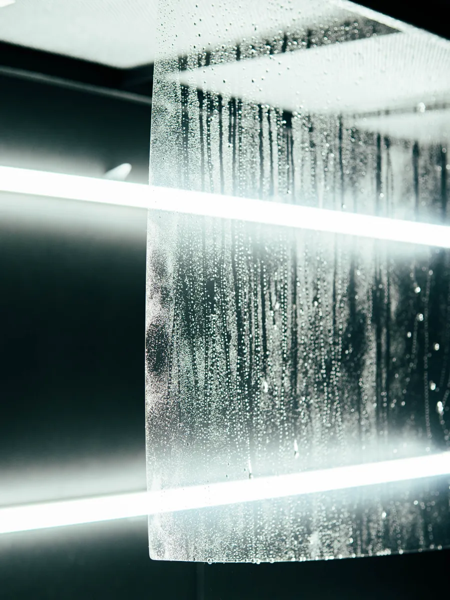 Close-up of a wet sheet of clear windshield protection film with its thick edge in focus, hanging in an install bay